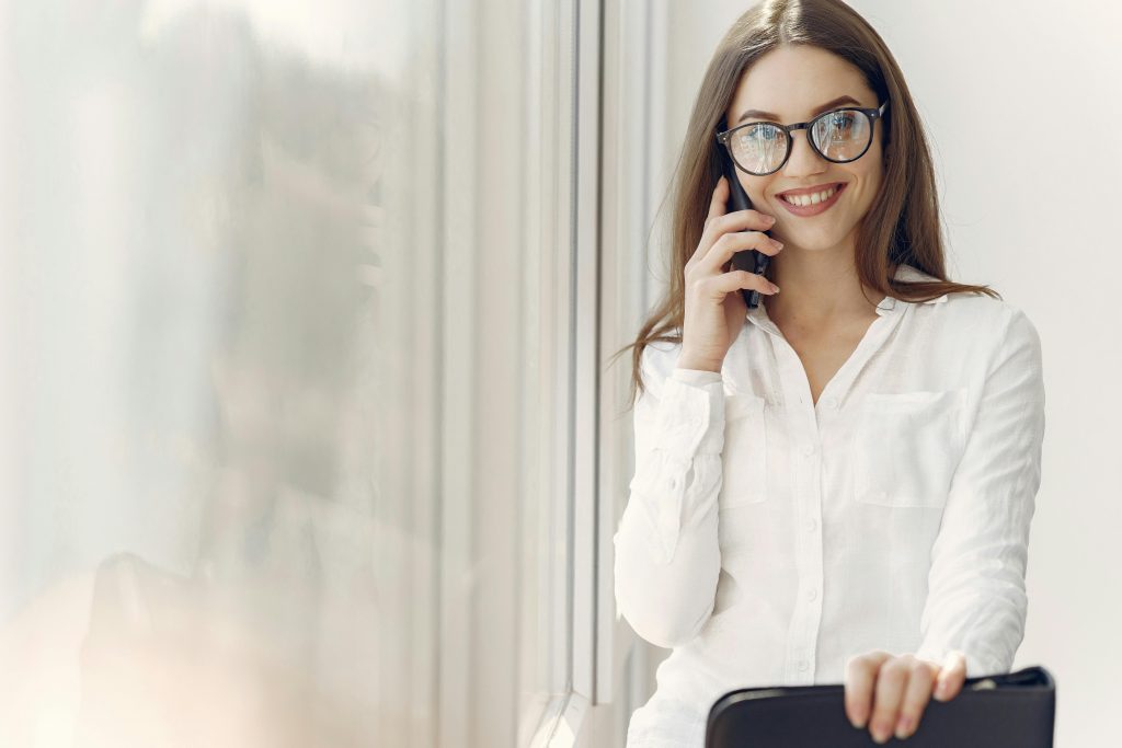 Cheerful woman in glasses talking on smartphone by a window indoors.